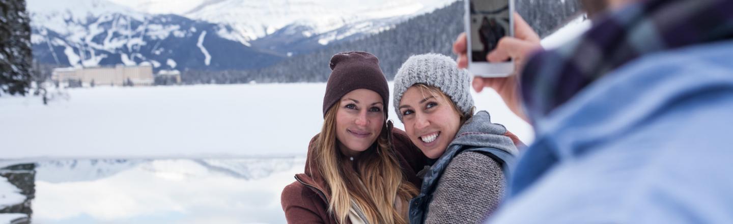 Two women wearing winter hats and scarves hugging while a man takes their photo with a smart phone. A frozen lake is seen in the background.