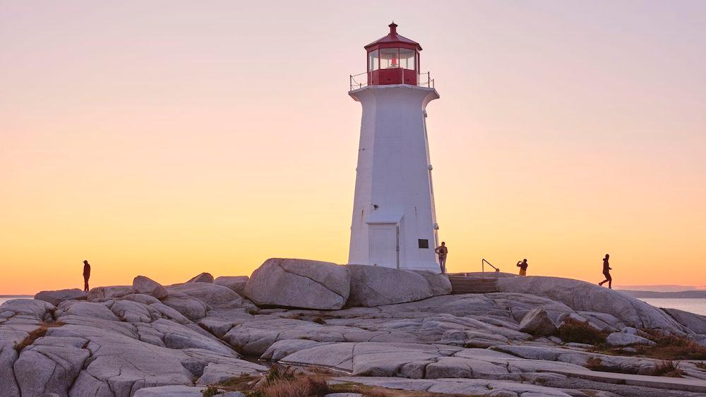 A few people walk along the large rocks in front of the Peggy's Cove lighthouse at sunset.